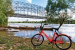 A bright red bike sits on a riverbank strewn with brown leaves and some visible green grass. Large logs can bee see in the shallow river water. In the distance there is a large, metal bridge spanning the river.