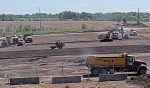 Construction equipment can be seen clearing a large flat area of dirt, with green trees and sky in the far distance